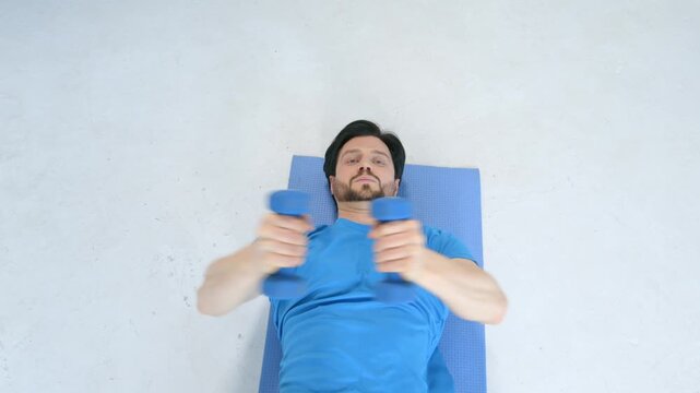 Overhead view of active fit man in sportswear lying on yoga mat, practicing dumbbell chest fly exercise, increasing shoulder and core strength while working out at home.