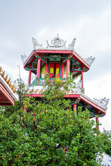 Fototapeta premium A bright Chinese-style pavilion rises above lush green trees, its red pillars and ornate roof decorations standing out against the pale sky.
