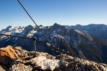 Mountain summit view with rocky foreground and cable anchor, showcasing snow-capped peaks and clear blue sky in the Alps region during daylight