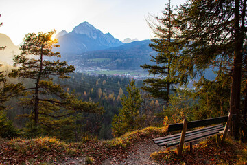 Scenic mountain view at sunset with a wooden bench overlooking a valley surrounded by trees and distant peaks in a tranquil natural setting