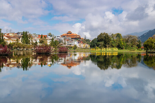 Scenic view of Sa Pa Lake with the city skyline and Fansipan peak in the background, taken in Sa Pa, Lao Cai, Vietnam. The lake reflects the sky and city.