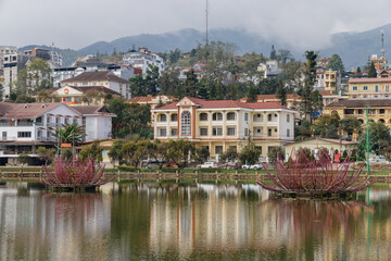 Fototapeta premium Scenic view of Sa Pa Lake with lotus sculptures, reflecting the town's architecture, in Vietnam. A tranquil scene showcasing local art and urban landscape.