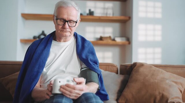 Elderly man using modern monitor of blood pressure for home use, medicine. Senior male person sitting on couch in living room, sick man feeling bad, hypertension and seasonal cold, stay healthy