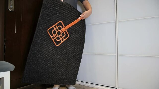 Detail view of person standing indoors, vigorously beating a large dark entrance mat with an orange plastic rug beater. Home cleaning activity for floor covering.
