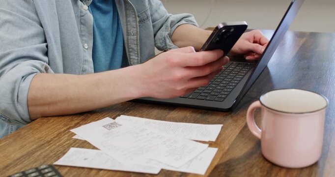 Man scrolls internet via smartphone as sits at desk with laptop and bills near tea mug closeup. Male entrepreneur processes monthly expenses working from home