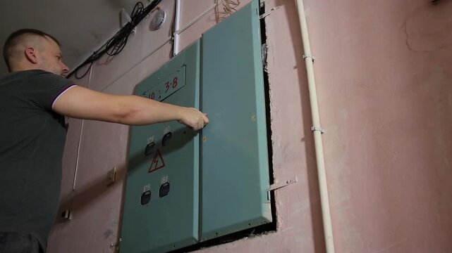 Close up view of adult man hand closing metal cover of industrial electrical panel with visible warning sign and meters, power supply system management and inspection by worker on building utility wal