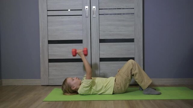 Child lies on green fitness mat on wooden floor, lifting small red weights above chest. Boy engaged in strength training, developing muscles and promoting healthy lifestyle indoors.