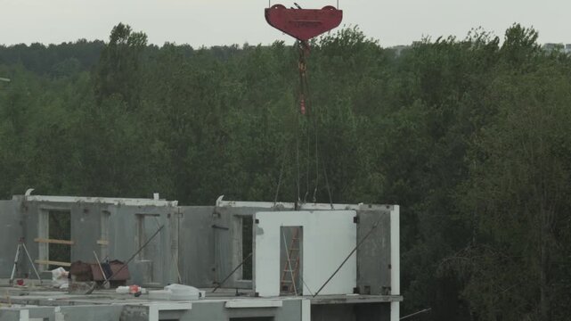 Industrial construction site view with large red crane hook lowering precast concrete wall panel onto partially completed residential building frame against forest backdrop urban development