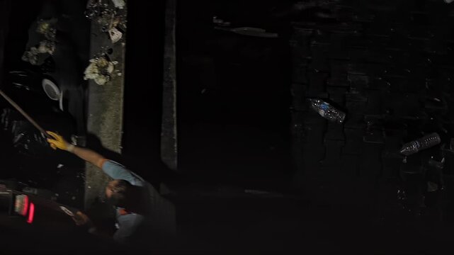 One person sanitation worker uses stick to push household garbage from street side bin into rear compartment of large waste collection vehicle during darkness city street.