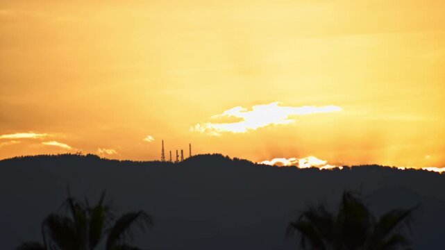 Dramatic morning light bathes atmosphere in warm orange and yellow hues as sun ascends behind dark hills. Towering antennae punctuate horizon, while blurred palm fronds offer tropical feel to dawn lan