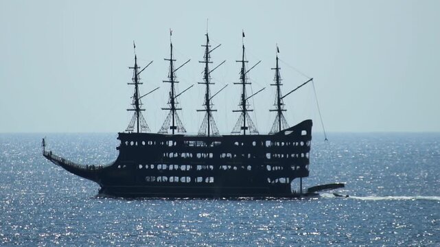 Dramatic silhouette of large multi-masted sailing ship on vast expanse of blue sea. Sunlight reflects on water. Ship appears dark against light sky, evoking journey and exploration. Slow motion x4