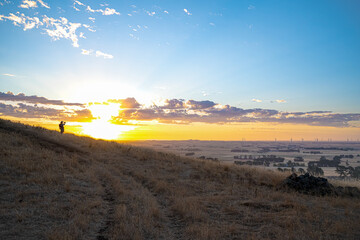 Fototapeta premium Landscape view of a female hiker watching and recording a golden sunset from the top of a hill overlooking rural Victoria, Mount Noorat, Victoria, Australia