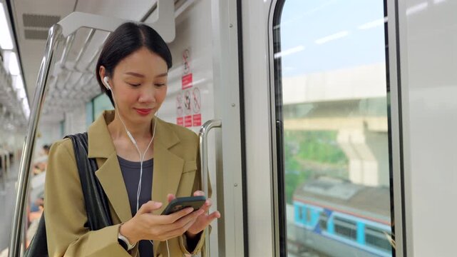 A modern Asian woman starts her daily morning routine with a productive commute on the city train. She is staying connected using her smartphone and earphones while enjoying the urban view.