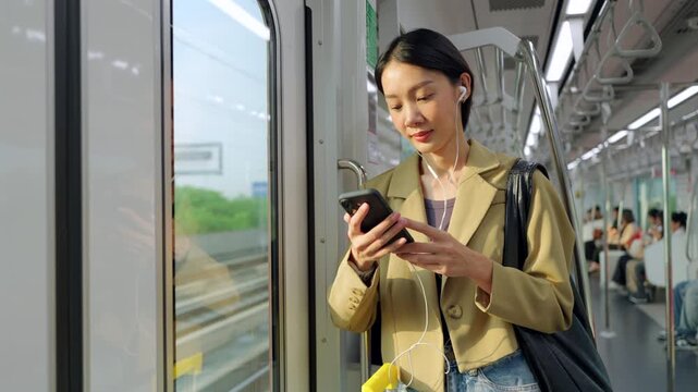 A modern Asian woman starts her daily morning routine with a productive commute on the city train. She is staying connected using her smartphone and earphones while enjoying the urban view.
