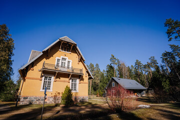 Obraz premium Two-story yellow house with balcony and gray roof surrounded by trees and grass under a clear blue sky in a rural setting