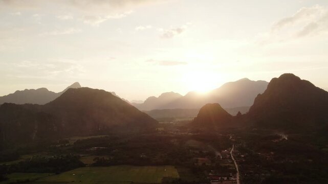 Flying airborne high altitude sideward panning moving view of mountainous landscape during sunset with paddy fields, small village in the foreground and paragliders and clear skies in the background.