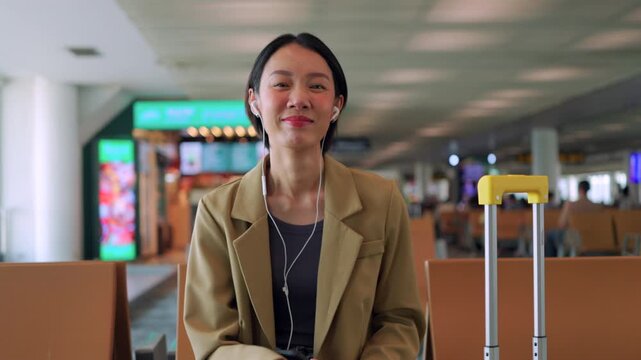 Happy Asian woman traveler waiting at the airport terminal, holding a passport and boarding pass while giving a thumbs up next to her yellow suitcase. Concept of journey and solo travel.