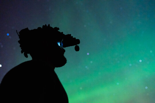 Tactical silhouette of a man in night vision goggles observing vibrant green Aurora Borealis in Greenland during night operation