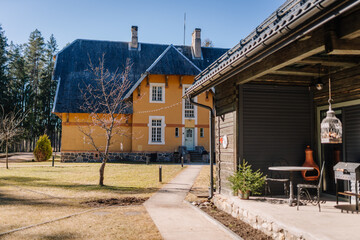 Naklejka premium Two-story yellow house with a steep roof and wooden accents, surrounded by a grassy yard and trees, with a stone pathway leading to a nearby wooden structure