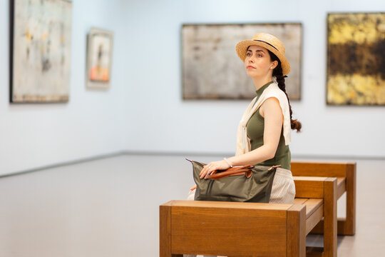 Young elegantly dressed woman in a straw hat admiring the exhibition of paintings in the gallery, sitting on bench. Contemporary art and cultural education concept