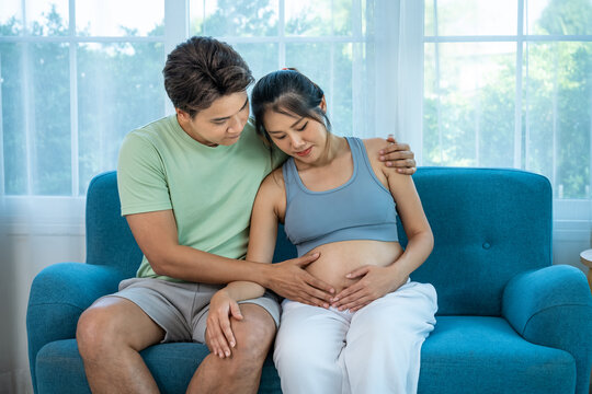 Asian expecting parents sharing tender moment, touching baby belly at home