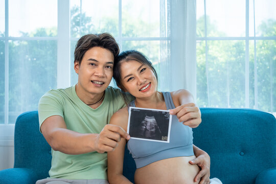 Joyful Asian couple sharing baby ultrasound, tender pregnancy moment at home, looking at camera