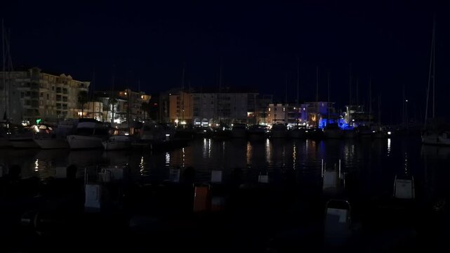 Handheld evening view on the dock in the city Frejus, France with street lamps and yachts parked, gentle waves and lights in the background