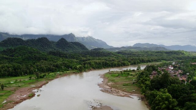 Sideward flying high altitude airborne panning view of a narrow river and it coastline with large moutain formations in the bac kground, in overcast rainy weather.