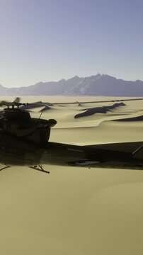 A helicopter hovers above golden sand dunes as the sun sets on the horizon. Mountains are visible in the background, creating a striking view at dusk.