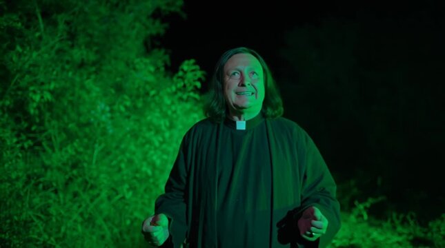 Mysterious priest with arms open wide is preaching under an eerie green light at night. Cassock