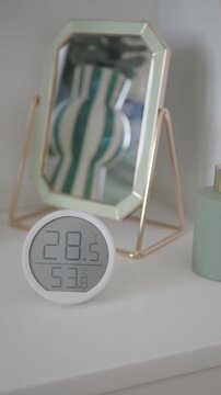 Digital thermometer and humidity sensor on white shelf with decorative mirror and reed diffuser
