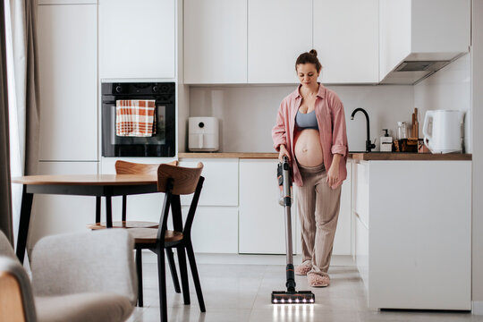 Pregnant woman cleaning floor with cordless vacuum in modern kitchen