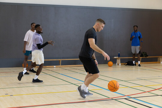 Four men dribbling basketballs on hardwood gym court, bench with bags and water bottle