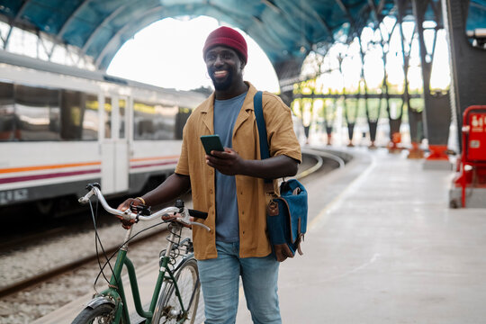 Smiling man with bicycle at train station using mobile phone