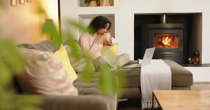 Nonbinary adult settling on sofa by fireplace, opening laptop, typing while sipping mug, copy space