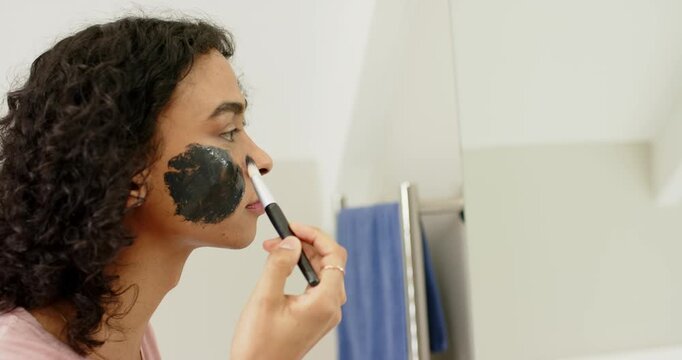 Dipping brush into charcoal mask jar, non-binary person applying across cheek at sink, copy space