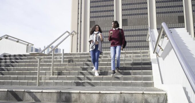 Diverse friends descending staircase at office sipping coffee and chatting and checking smartphone