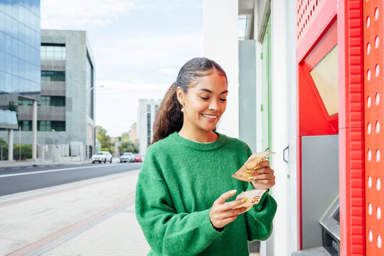 Smiling woman withdrawing euro cash at ATM in city street