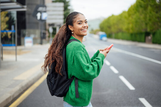 Smiling student with backpack and smartphone walking on urban street
