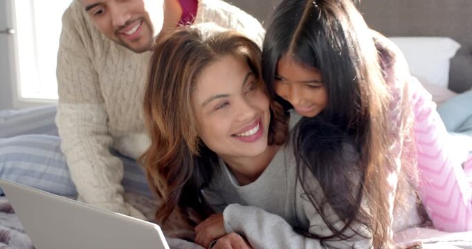 Diverse family on bed scrolling laptop when child climbing onto back and father kneeling laughing