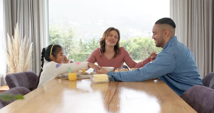 Hispanic family holding hands praying before serving spaghetti, eating at table by garden window