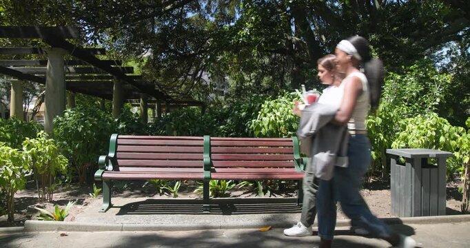 African American women entering left carrying cups, bag, sitting on park bench sharing snack
