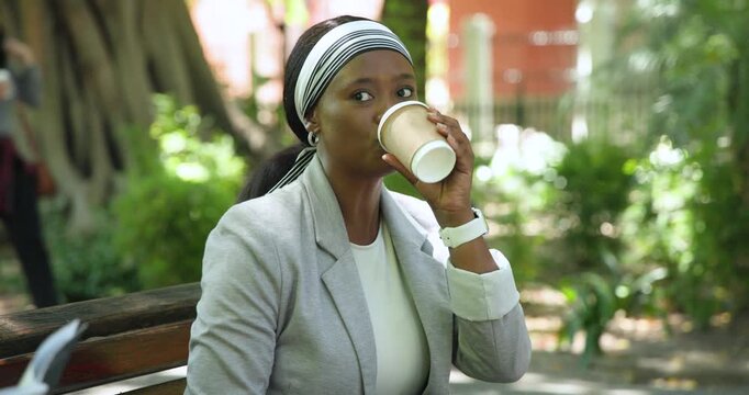 Adult African woman sitting on wooden bench in park, sipping coffee and scrolling phone on break