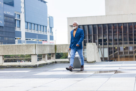 Senior african american man wearing blazer walking on tiled urban plaza and rolling suitcase