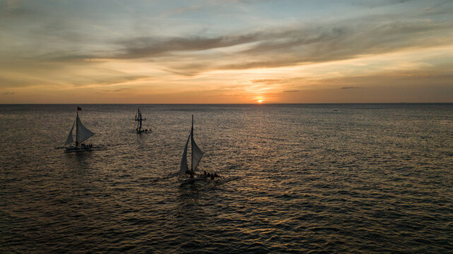 Aerial view of traditional Paraw sailboats sailing on the calm ocean at sunset in Boracay, Philippines. A beautiful tropical seascape during golden hour with vibrant orange sky