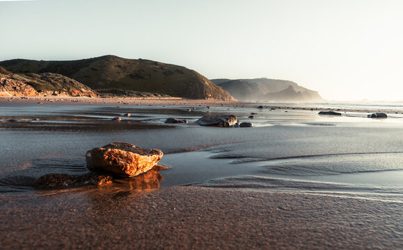 View of sun-kissed, wet sand glistens, reflecting the soft light as waves gently lap the shore under the distant hills, Portimao, Faro District, Portugal.