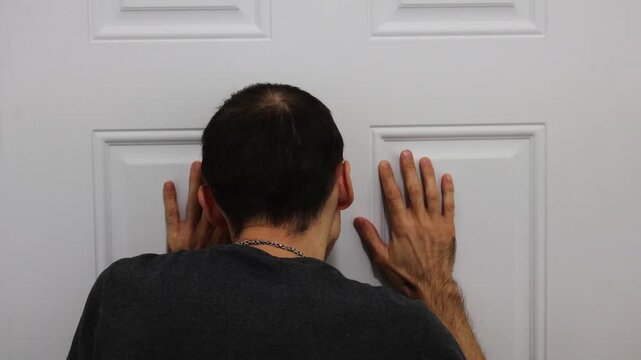 Man leaning on door with both hands while peeking through peephole indoors
