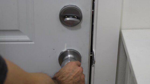 A hand moves back to the door knob after unlocking the deadbolt, preparing to fully open the door.
