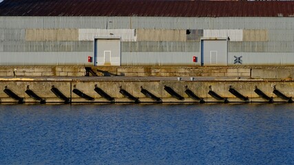 Naklejka premium Industrial harbor pier with black fenders and rhythmic long shadows on concrete wall. Corrugated metal warehouse background. Maritime logistics and port infrastructure. Sunset lighting.