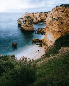 View of rugged golden cliffs meet the turquoise sea, framing a secluded beach where tiny figures stroll, under a soft sky, Portimao, Faro, Portugal.